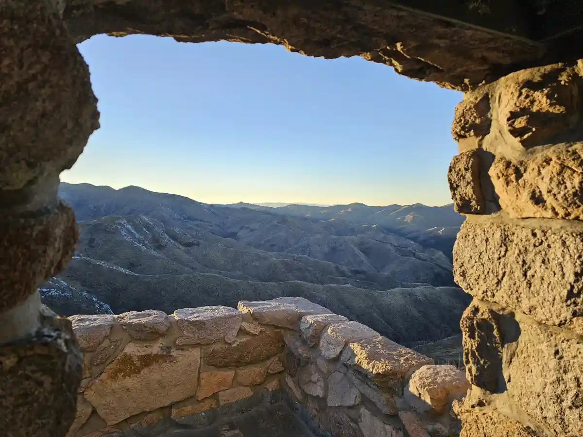 A scenic view of rolling mountains under a clear blue sky, framed by the rough, weathered stones of an old stone structure…
