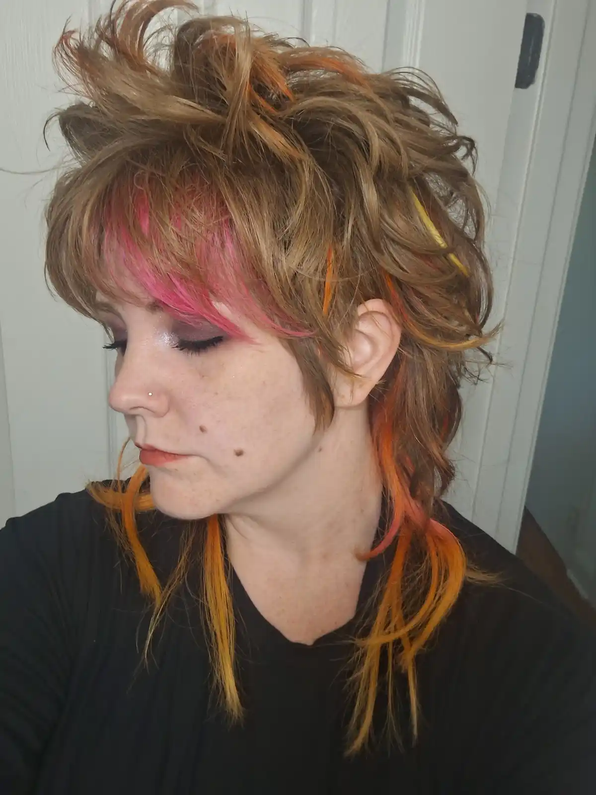 A person with messy, multi-colored hair featuring bright pink bangs, orange streaks, and yellow highlights, looking down…