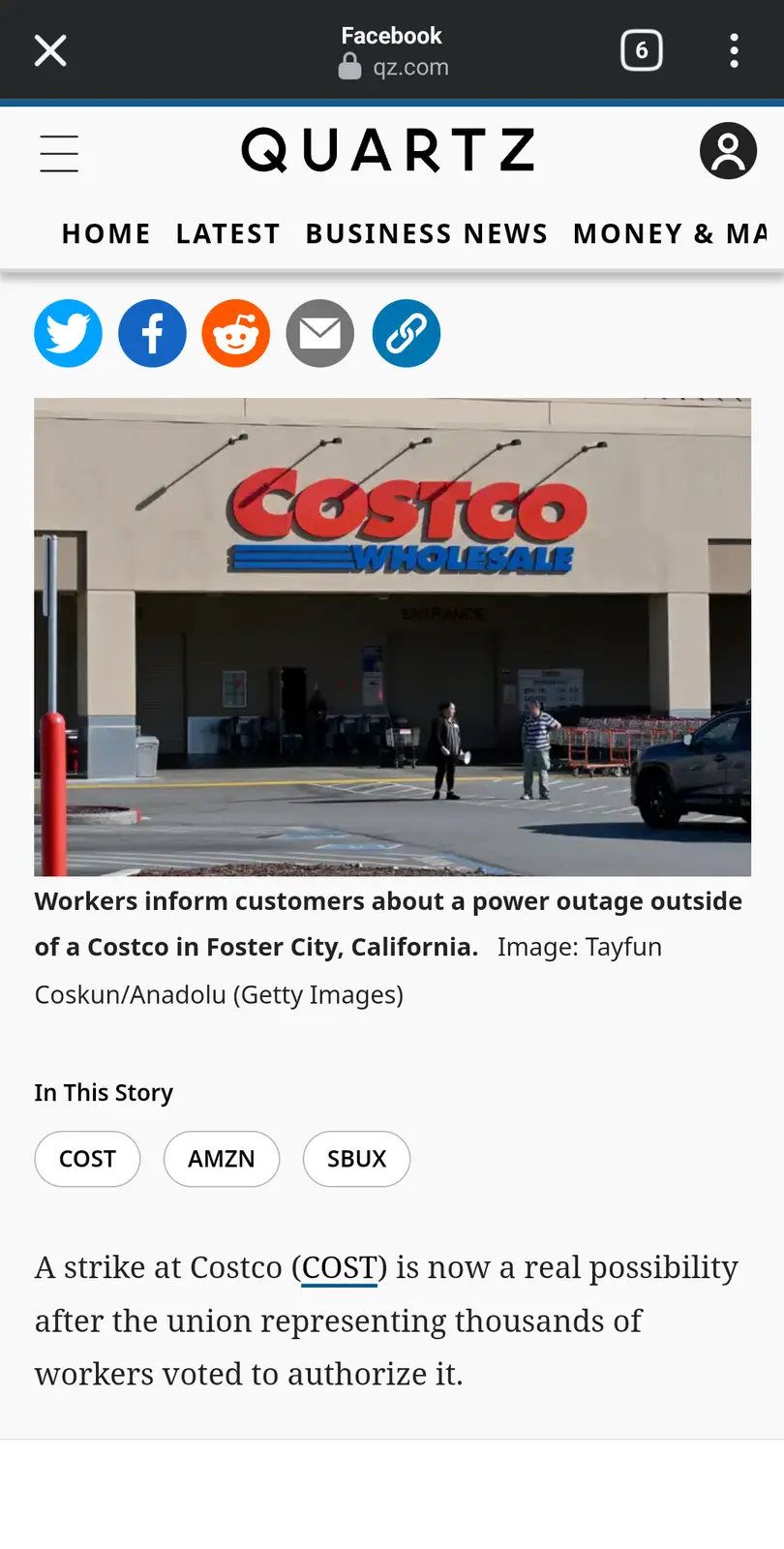 The image shows the exterior of a Costco Wholesale store with its large red and blue logo. Two workers stand in the parking…