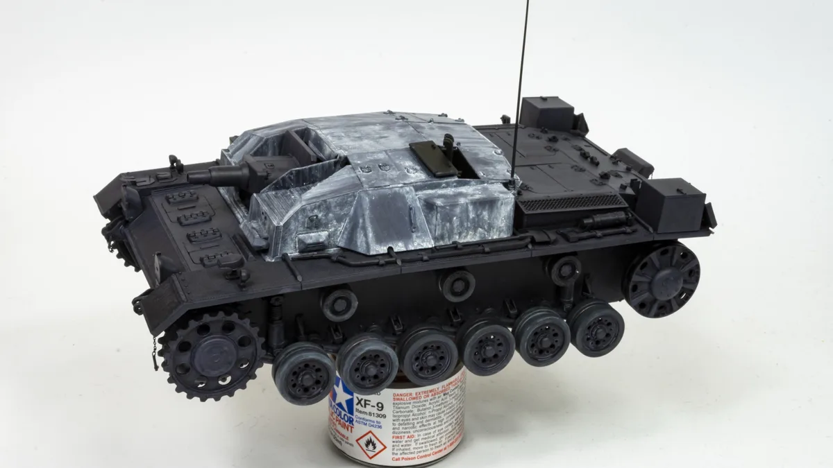 A detailed scale model of a German World War II tank, likely a StuG III, is shown resting on a can of Tamiya XF-9 acrylic…