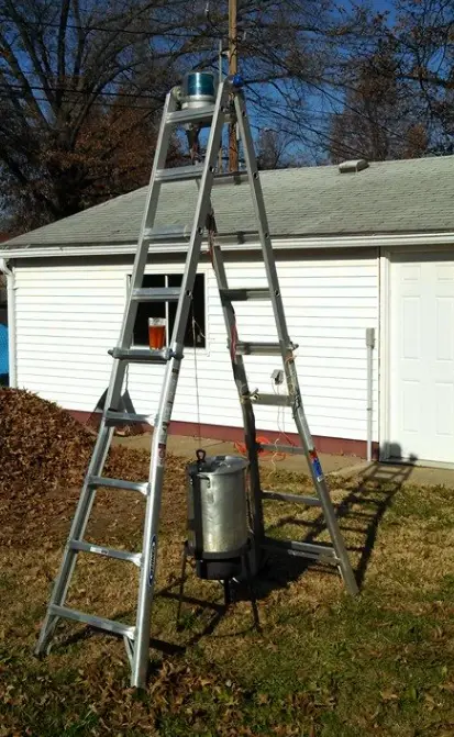 An outdoor photo shows a tall aluminum ladder leaning against a utility pole with a small container on top. At the base of…