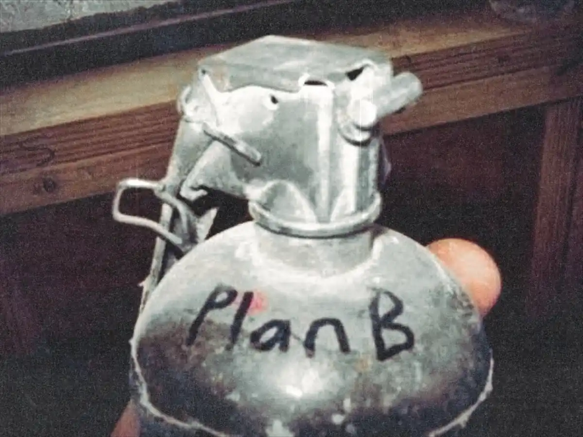 A close-up photo shows a hand holding a metallic hand grenade with the words 'Plan B' written in black marker on its body…