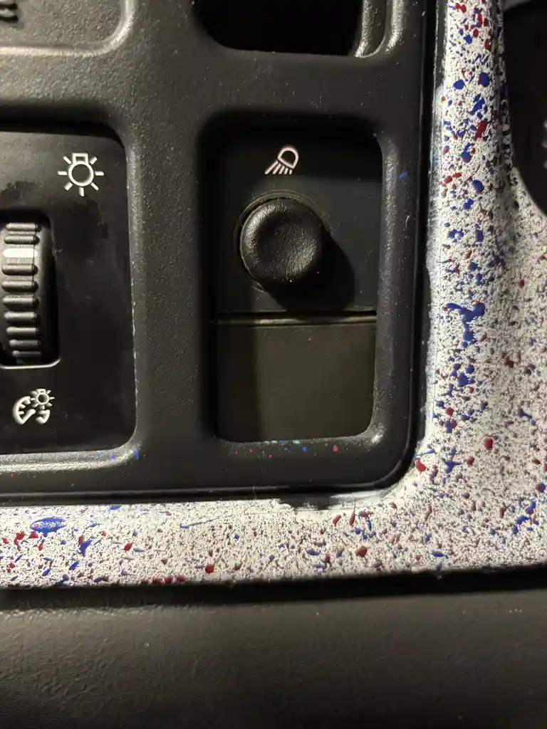 A close-up of a car's dashboard controls shows a black button with a windshield wiper icon, next to a light switch. The…
