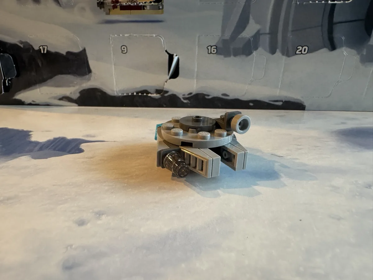 A small, gray Lego piece resembling a spaceship part sits on a white surface in front of a partially torn cardboard box with…