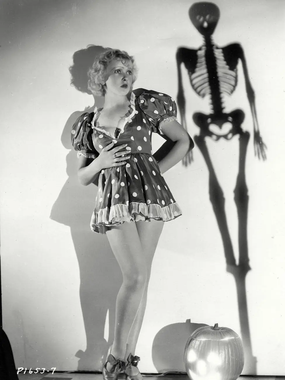 A vintage black-and-white photo shows a woman in a polka-dot dress looking terrified as a large skeleton shadow looms behind…