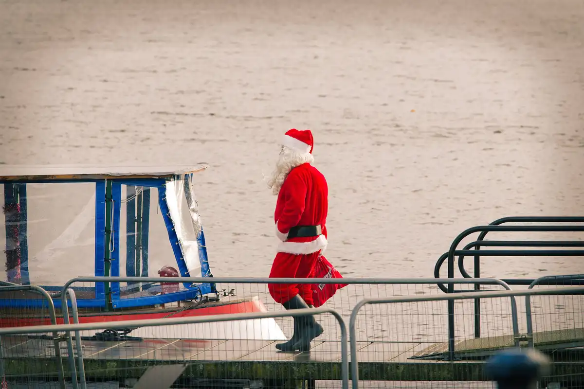 A person dressed as Santa Claus in a red suit with white trim and black boots walks away from a small boat docked at a pier…