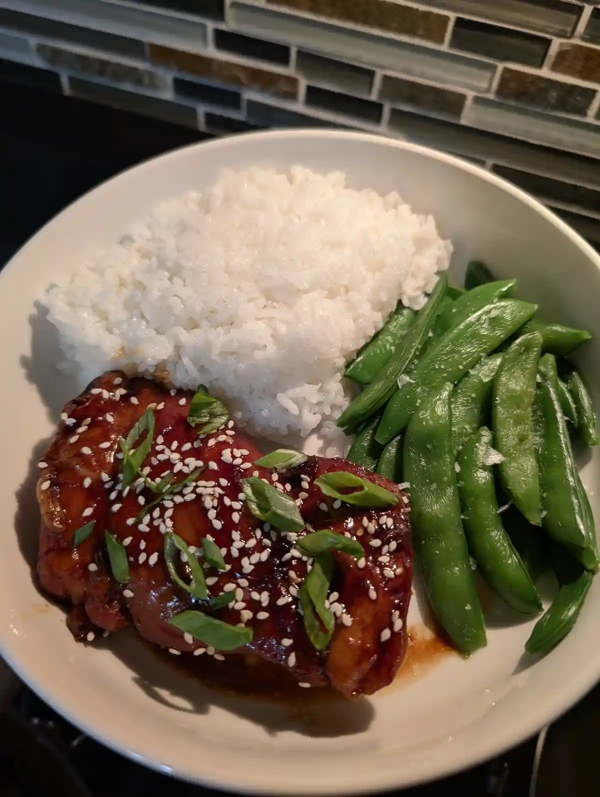 A white bowl contains a meal of glazed chicken topped with sesame seeds and green onions, served with white rice and steamed…