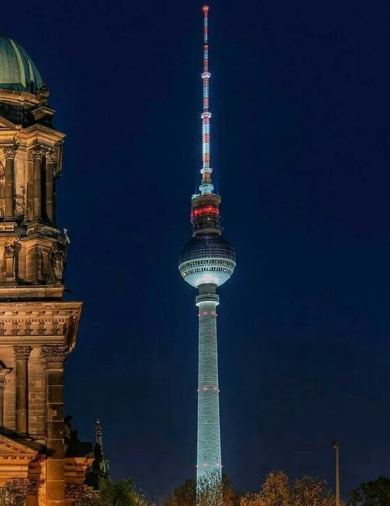 A nighttime photo of the Berlin TV Tower, illuminated with blue and red lights, standing next to the historic Berlin…