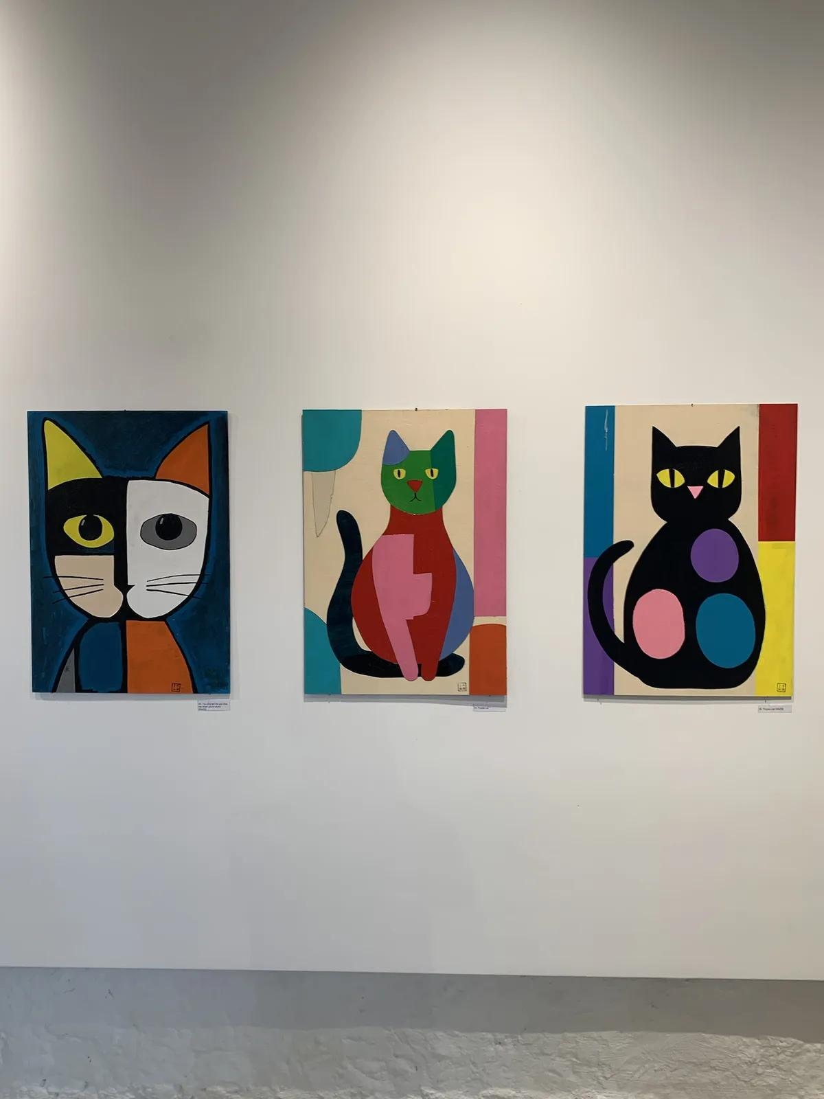 Three colorful, abstract cat paintings hang side-by-side on a white gallery wall. The left painting shows a cat split into…