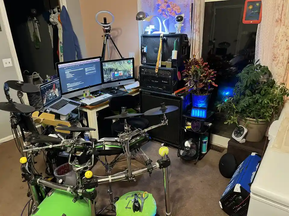 A cluttered room features an electronic drum set built directly onto a desk, with multiple monitors, a laptop, and audio…