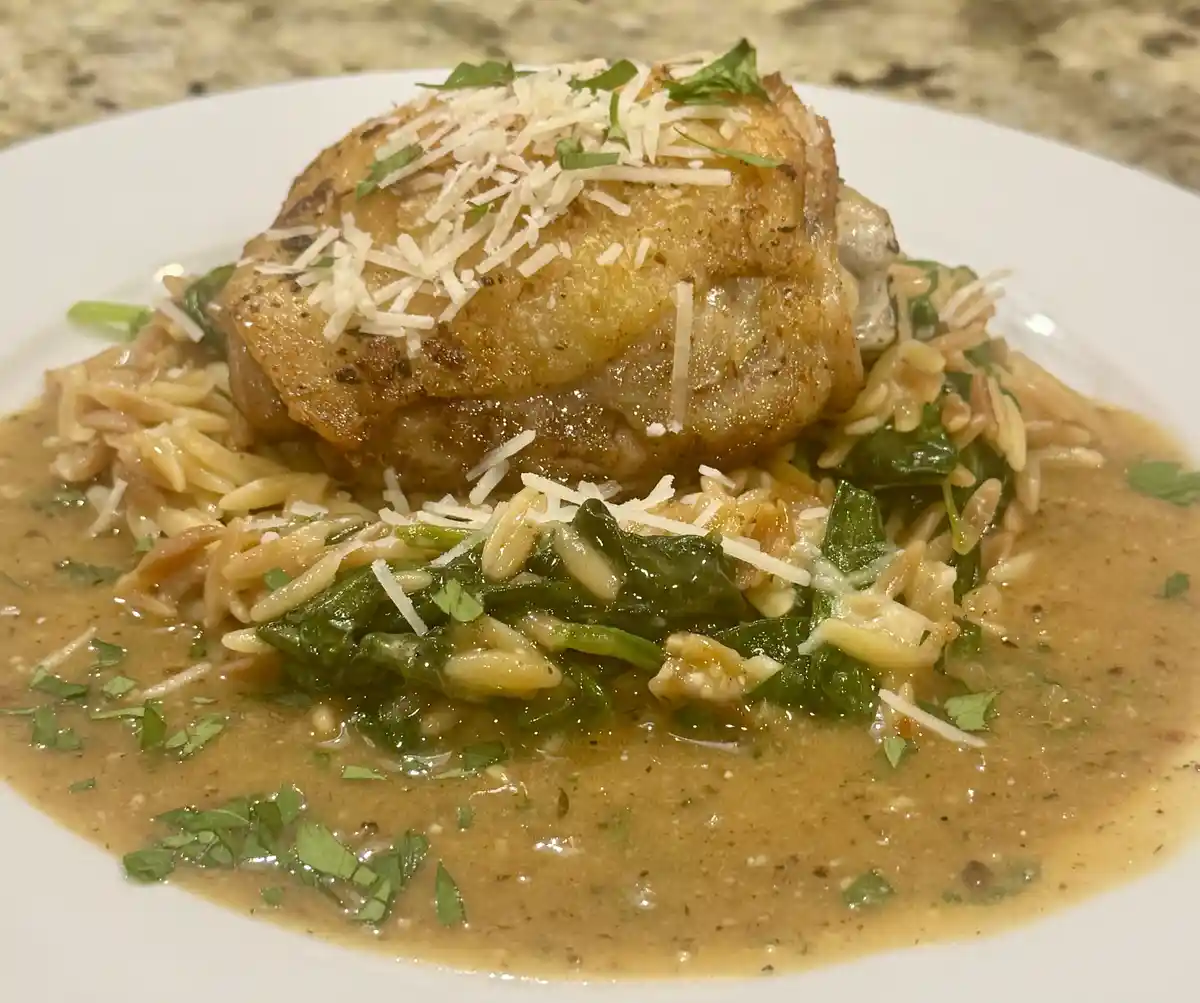 A close-up photo of a plated meal featuring a golden-brown chicken thigh atop a bed of orzo pasta mixed with wilted spinach…