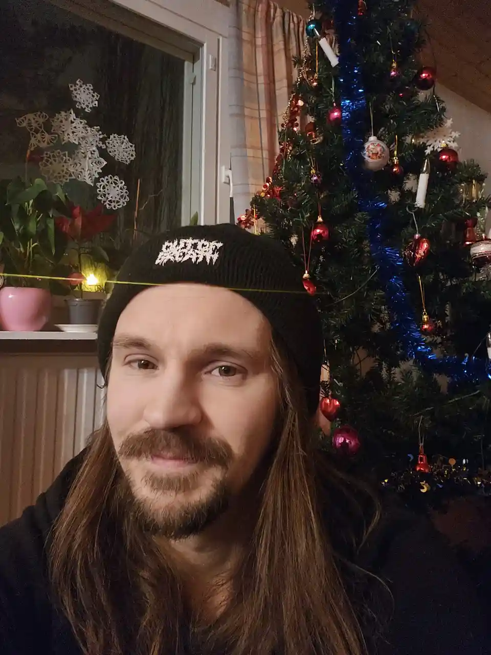 A man with long hair and a black beanie stands in front of a decorated Christmas tree with red ornaments and blue tinsel…