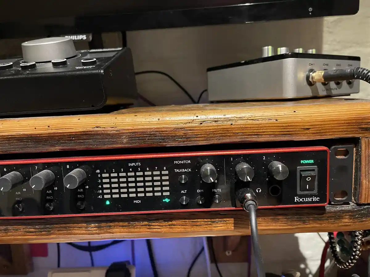 A close-up photo of a black and red Focusrite audio interface sitting on a wooden shelf, with various knobs, input levels…