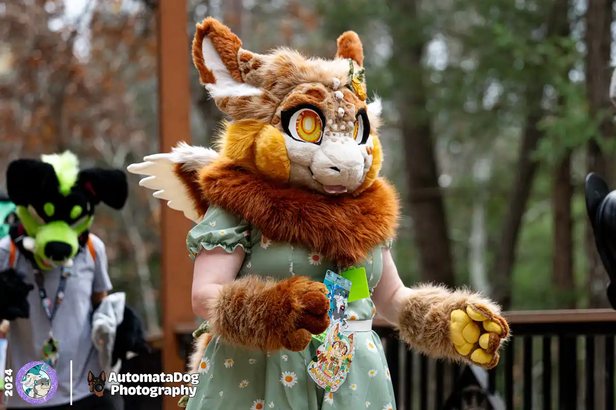 A person in a detailed brown and orange fursuit with large expressive eyes, white wings, and a green floral dress poses with…