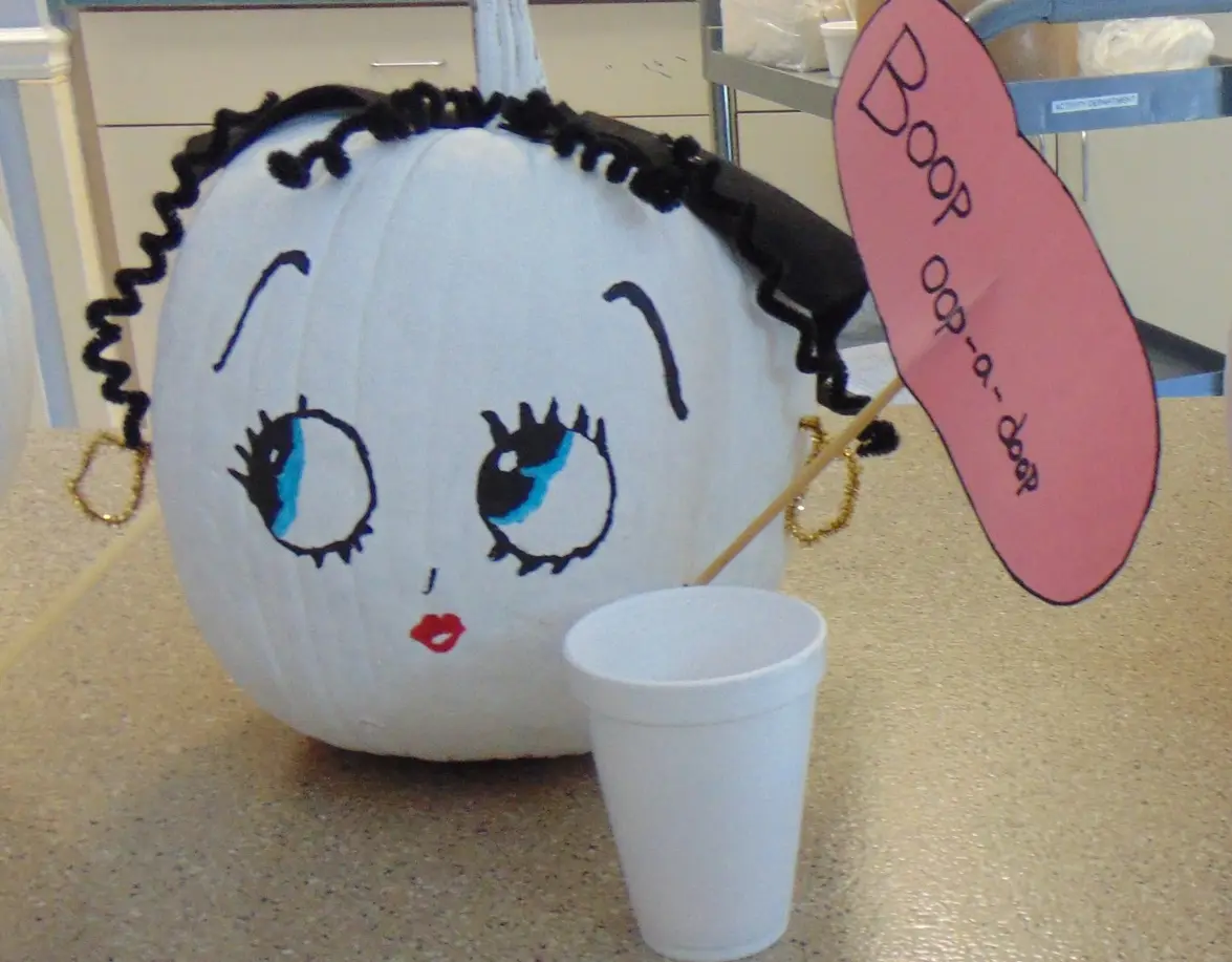 A white pumpkin decorated to resemble the cartoon character Betty Boop, featuring drawn-on blue eyes with lashes, red lips…
