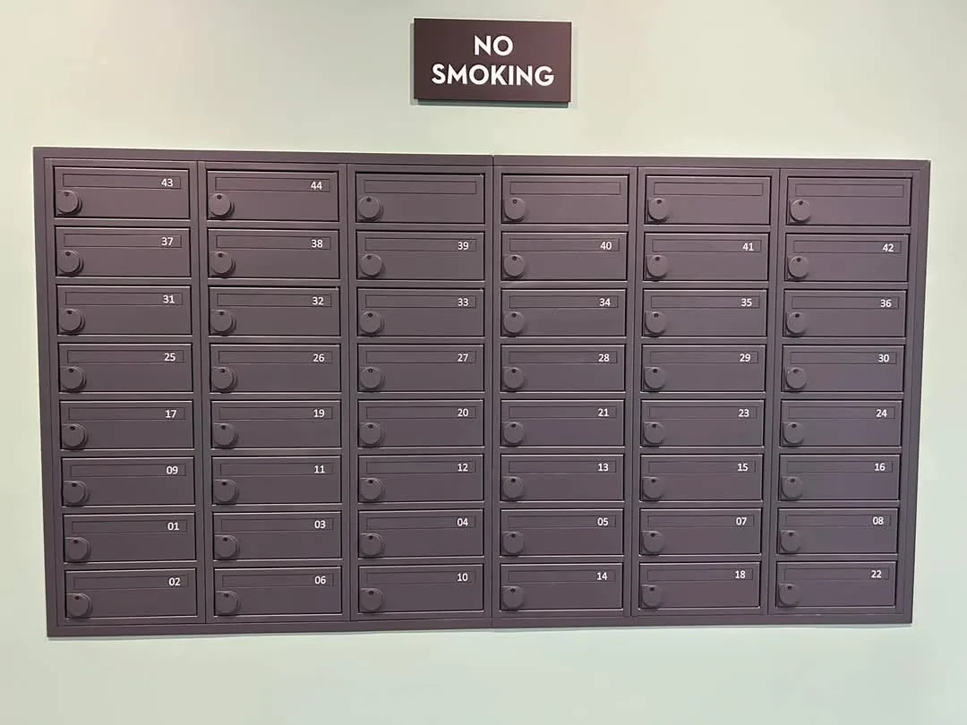 A wall-mounted array of numbered metal mailboxes, each with a small round handle, is shown under a brown sign that reads 'NO…