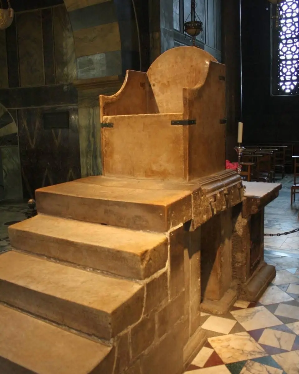 a large, ornate, stone throne-like chair sits on a raised platform with steps in what appears to be a church or cathedral…