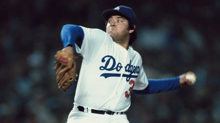 a baseball pitcher in a white Dodgers jersey with blue sleeves and number 3, mid-throw with a glove on one hand and a…