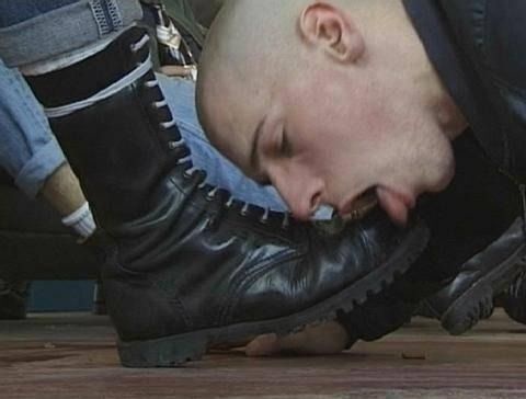 A bald man is kneeling and kissing the toe of a black leather boot worn by another person. The person wearing the boot has…