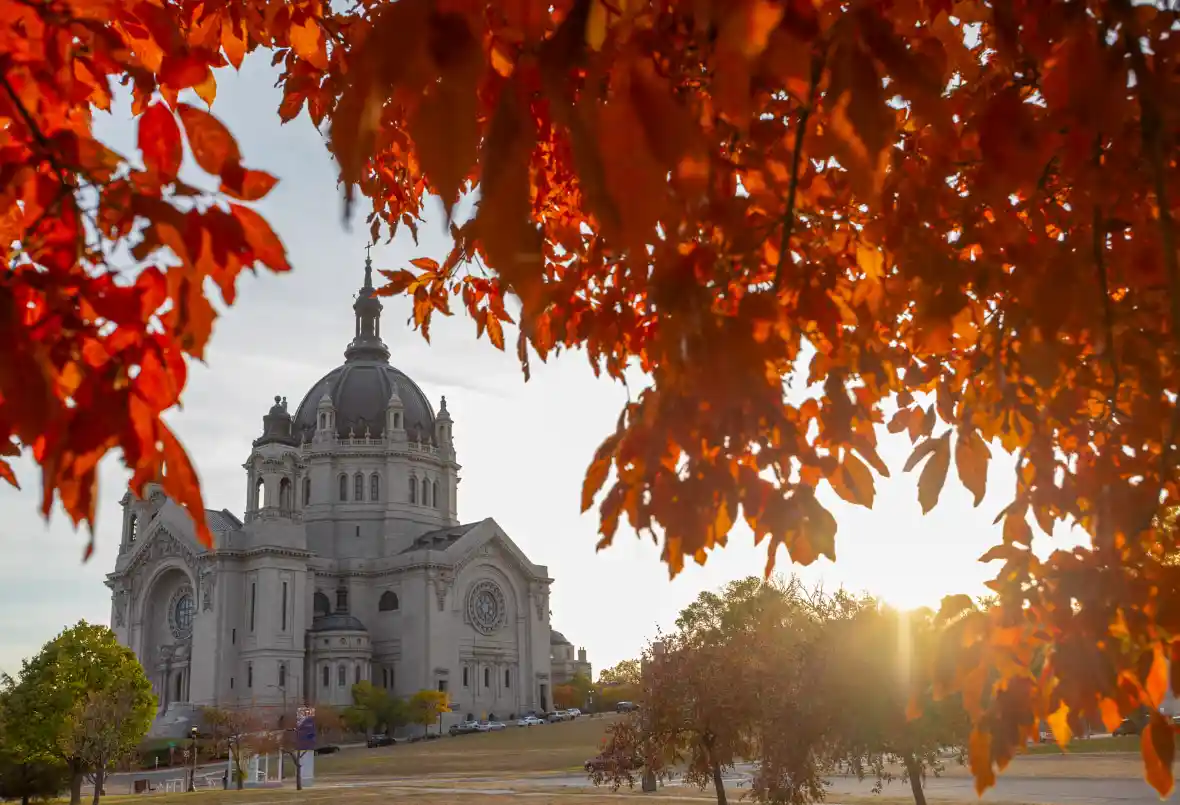 A scenic photograph of a large, ornate stone cathedral with a prominent dome, framed by vibrant orange and red autumn leaves…