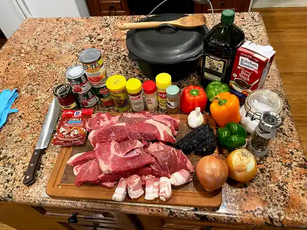 A kitchen counter is covered with ingredients for a large meal, including raw meat, bacon, bell peppers, onions, garlic…