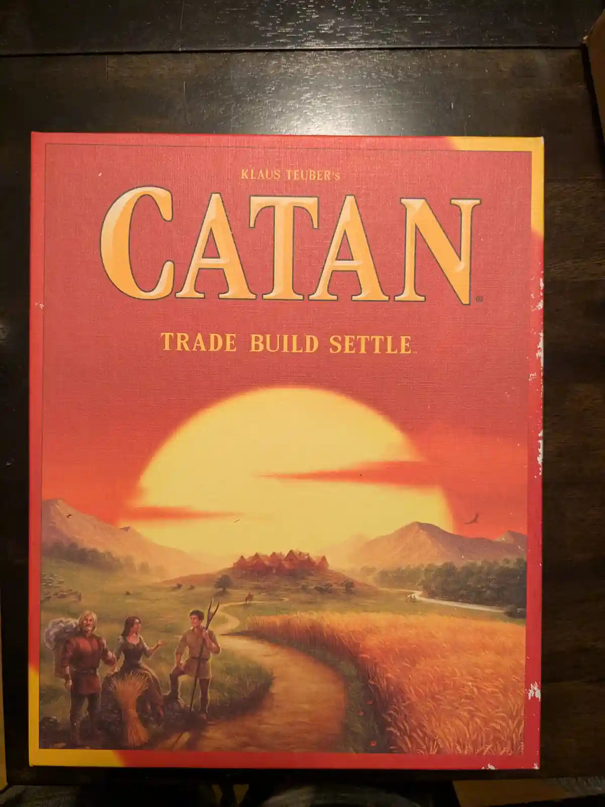 The image shows the front cover of the board game 'Catan' by Klaus Teuber, featuring a painted scene of three settlers on a…
