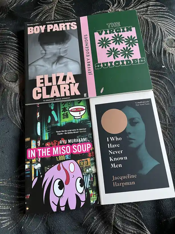 Four books are arranged on a dark, textured fabric background. Top left is 'Boy Parts' by Eliza Clark with a black-and-white…