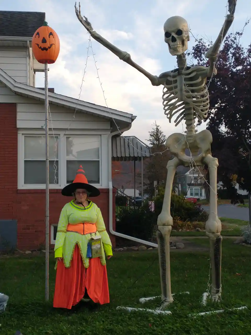 A person dressed as a witch in a bright green and orange costume stands next to a large outdoor skeleton decoration. The…