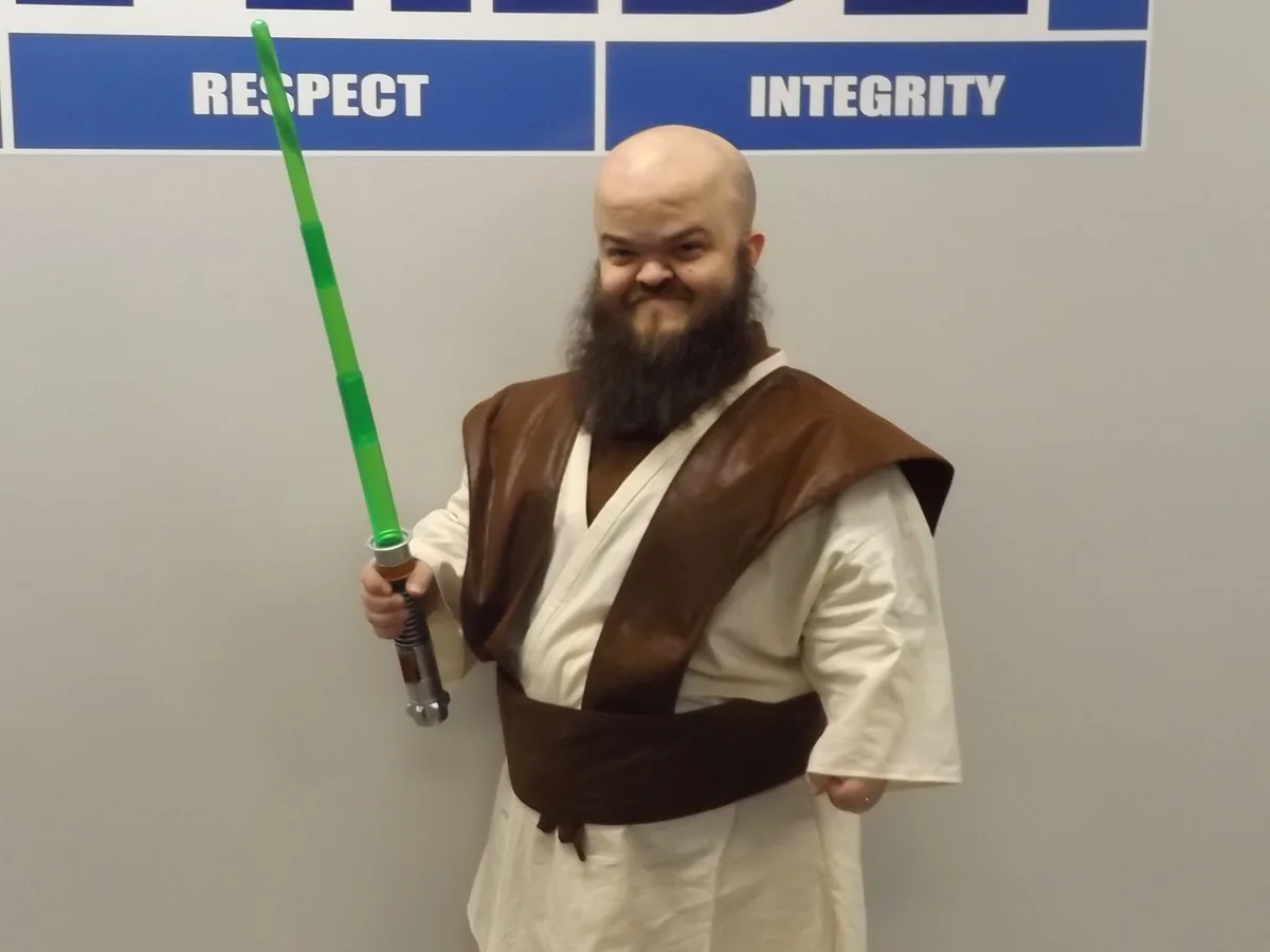 A bald man with a long beard dressed in a Jedi robe and holding a green lightsaber stands in front of a wall with blue signs…