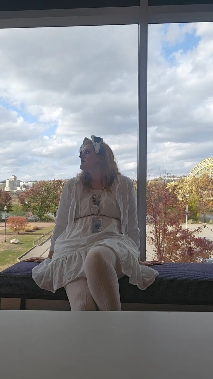 A woman in a white dress and white-and-black cow-print sunglasses sits on a bench indoors, looking upward with a pensive…
