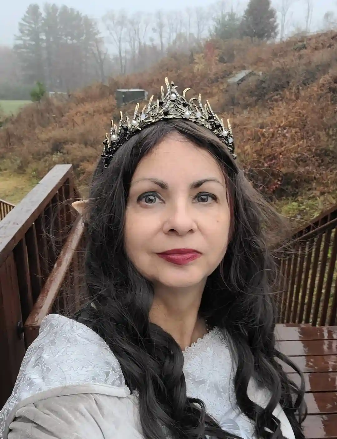 A woman with long dark hair, wearing a silver and black jeweled crown and a white lace top, stands on a wooden deck…
