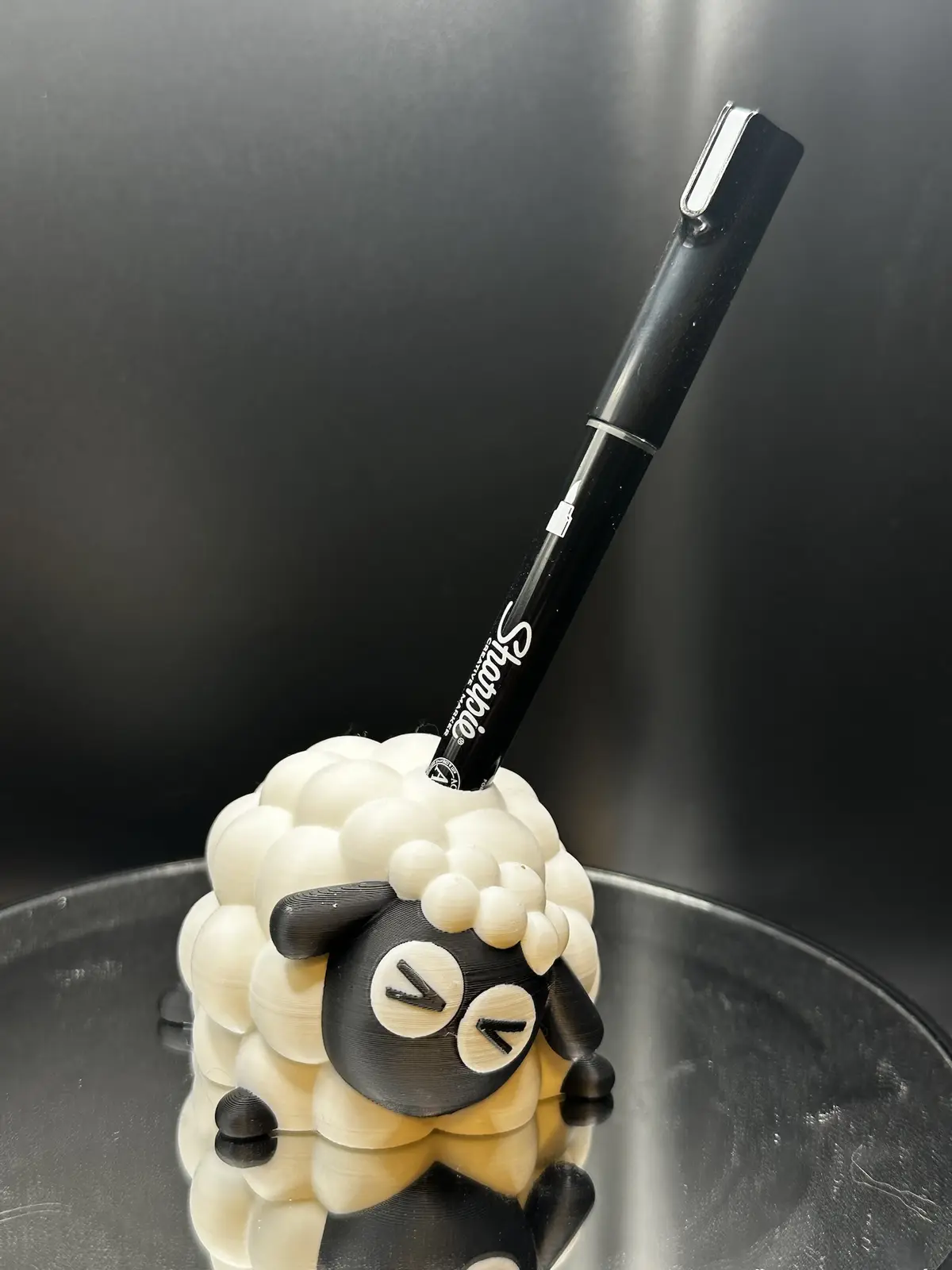 A black Sharpie marker is stuck upright in a 3D-printed sheep-shaped pen holder, which has a black face with white eyes and…