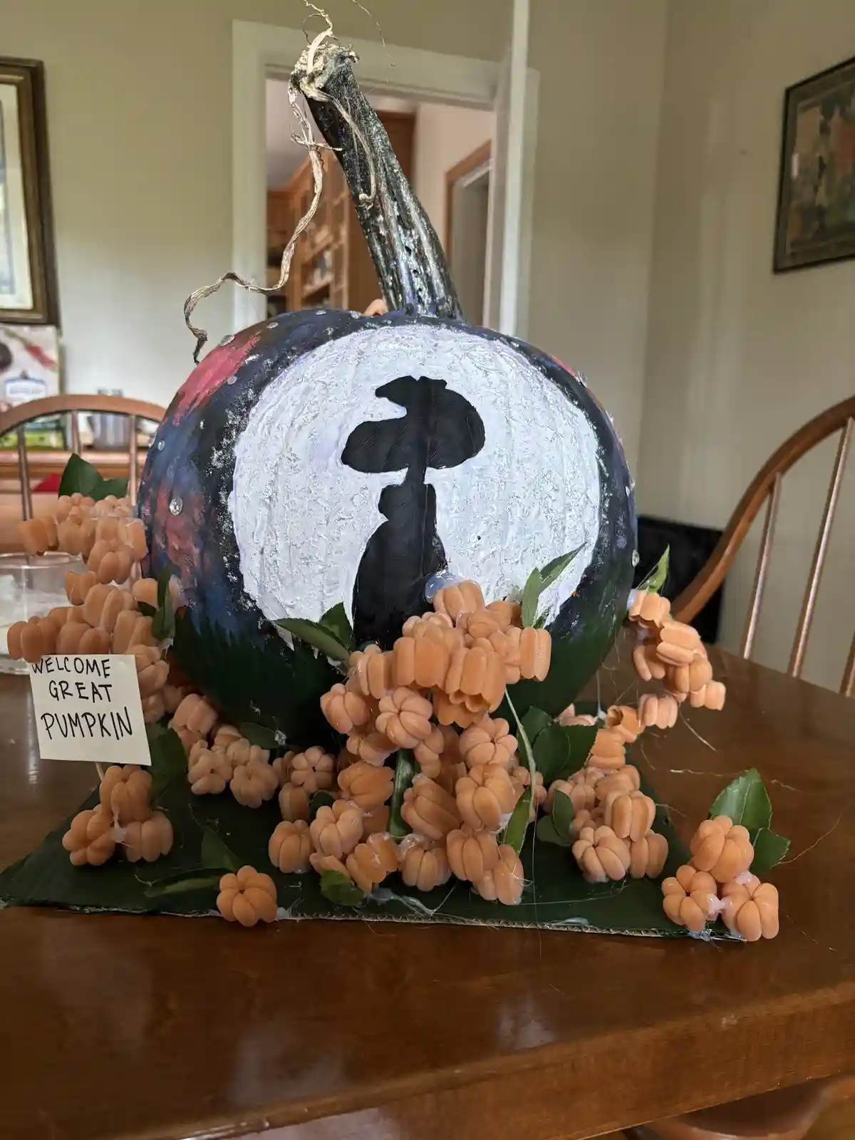 A large, decorated pumpkin sits on a wooden table, painted with a black silhouette of a figure against a white moon-like…