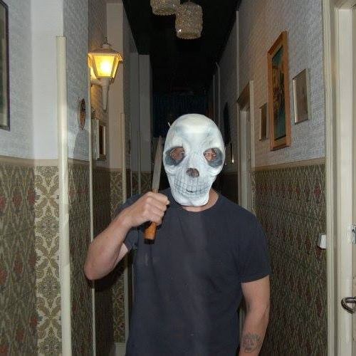 A person wearing a white skull mask and holding a knife stands in a dimly lit hallway with patterned wallpaper and a…