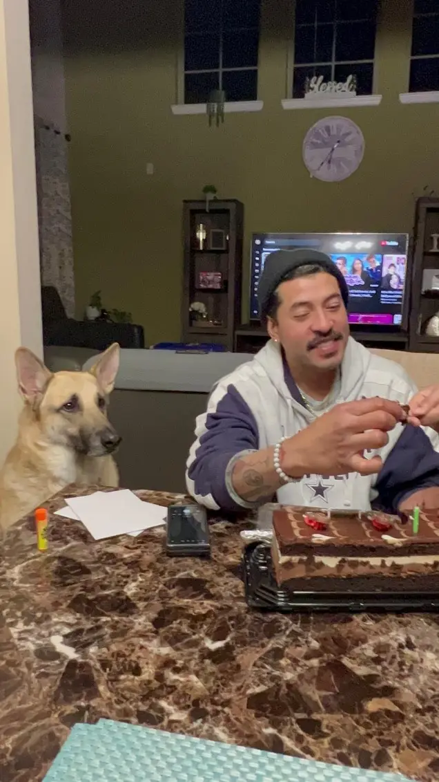 A man in a hoodie and beanie sits at a marble table with a chocolate cake and two lit candles, smiling as he holds a piece…