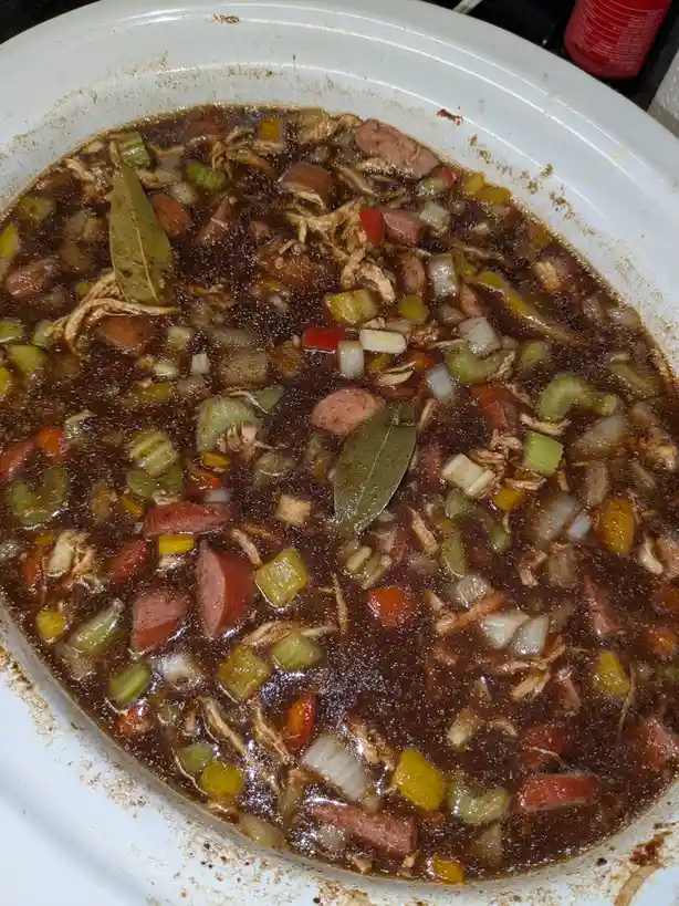 A close-up photo shows a white slow cooker filled with a dark, bubbling gumbo containing shredded chicken, chunks of…
