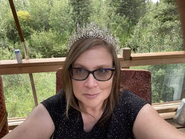 A woman with glasses and a tiara sits on a porch chair, looking directly at the camera with a neutral expression. Behind her…