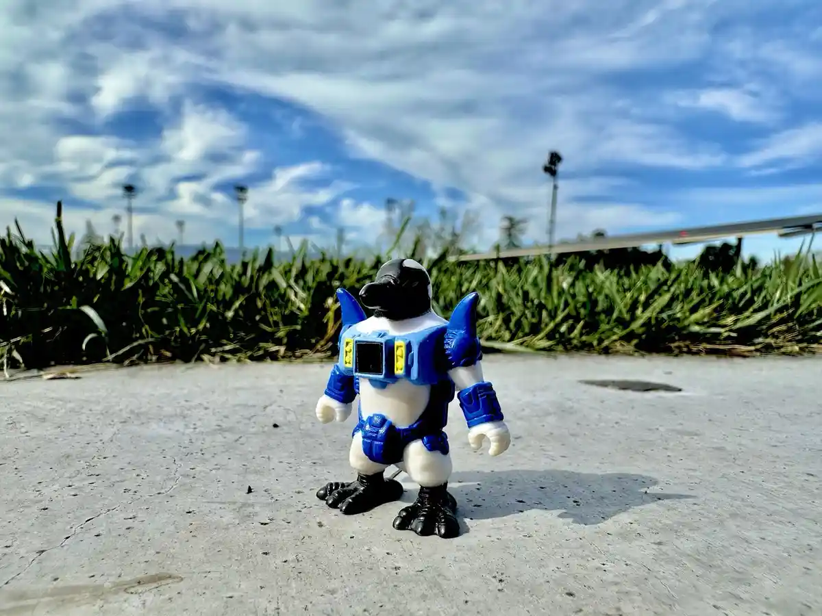A small plastic toy figure resembling a penguin in a blue and white armored suit stands on cracked concrete outdoors under a…