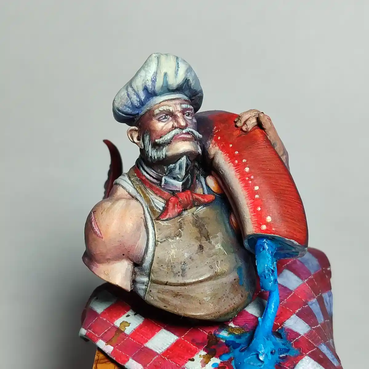 A painted miniature figurine of a muscular, bearded man with a chef's hat and apron, holding a giant red mushroom with blue…