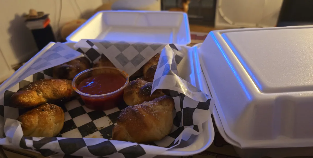A close-up photo shows a white styrofoam takeout container open to reveal a serving of golden-brown, seasoned breadsticks…