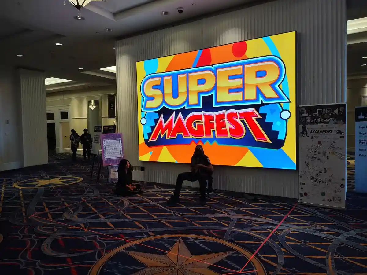 Two people sit on the floor in front of a large, colorful 'SUPER MAGFEST' sign at a convention hall, with a patterned carpet…