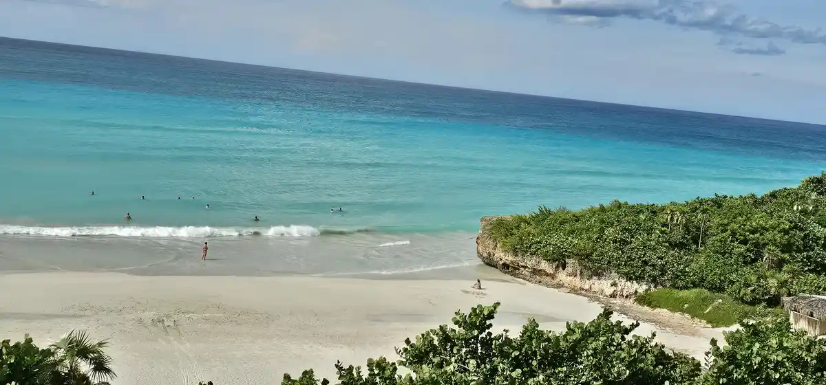 A wide-angle beach photo shows turquoise ocean water with several people swimming and one person standing in shallow water…
