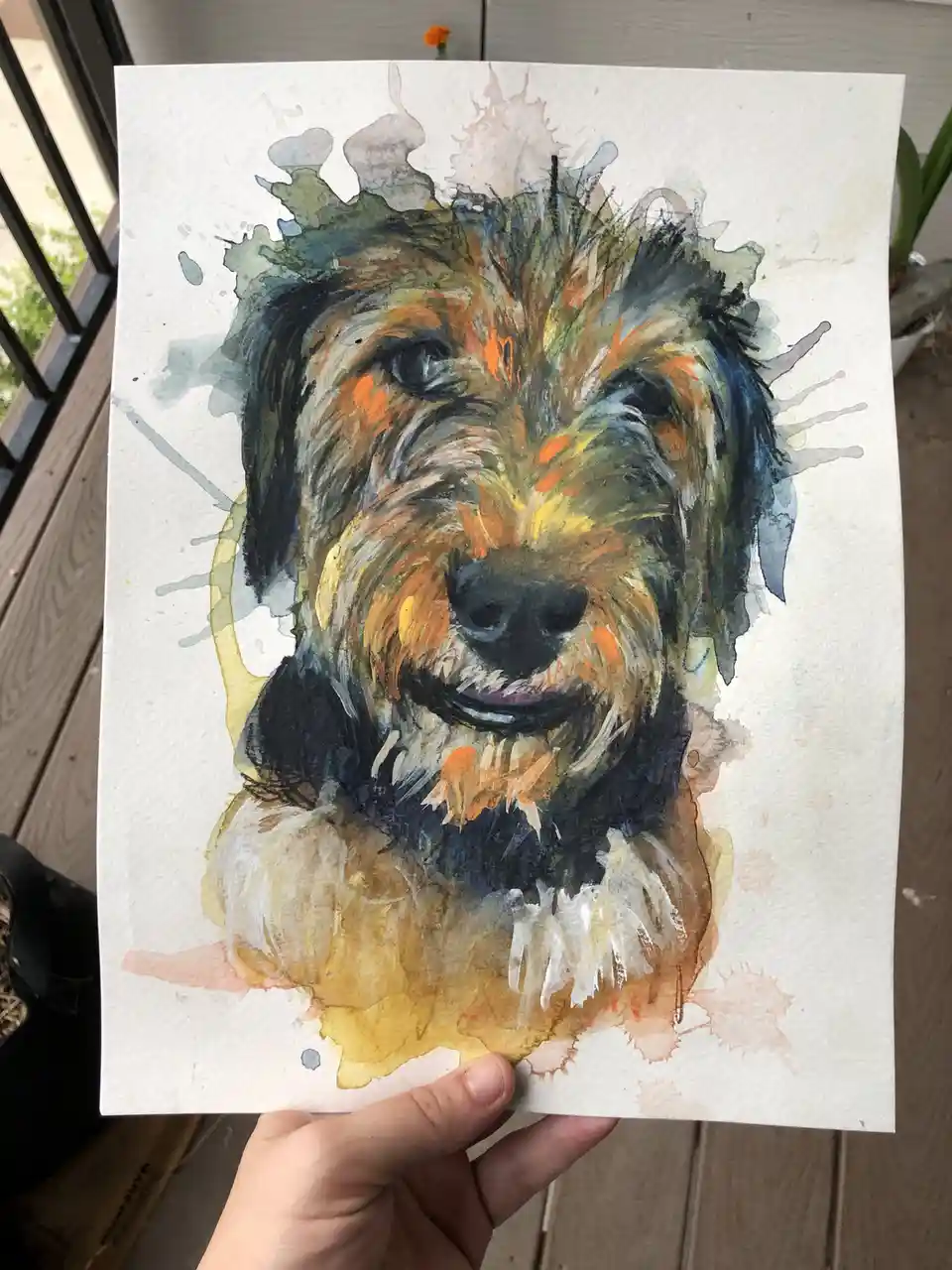 A hand holds up a watercolor painting of a scruffy, smiling dog with multicolored fur, surrounded by paint splatters on…