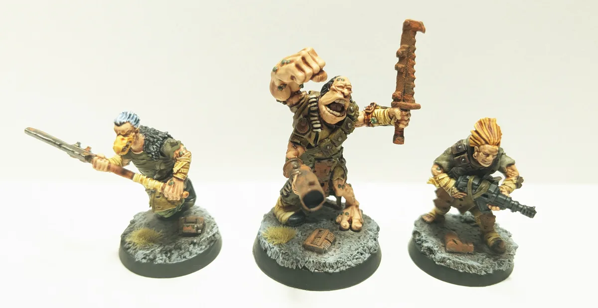 Three painted miniature figures, resembling grim, battle-worn warriors, stand on rocky bases against a plain white…