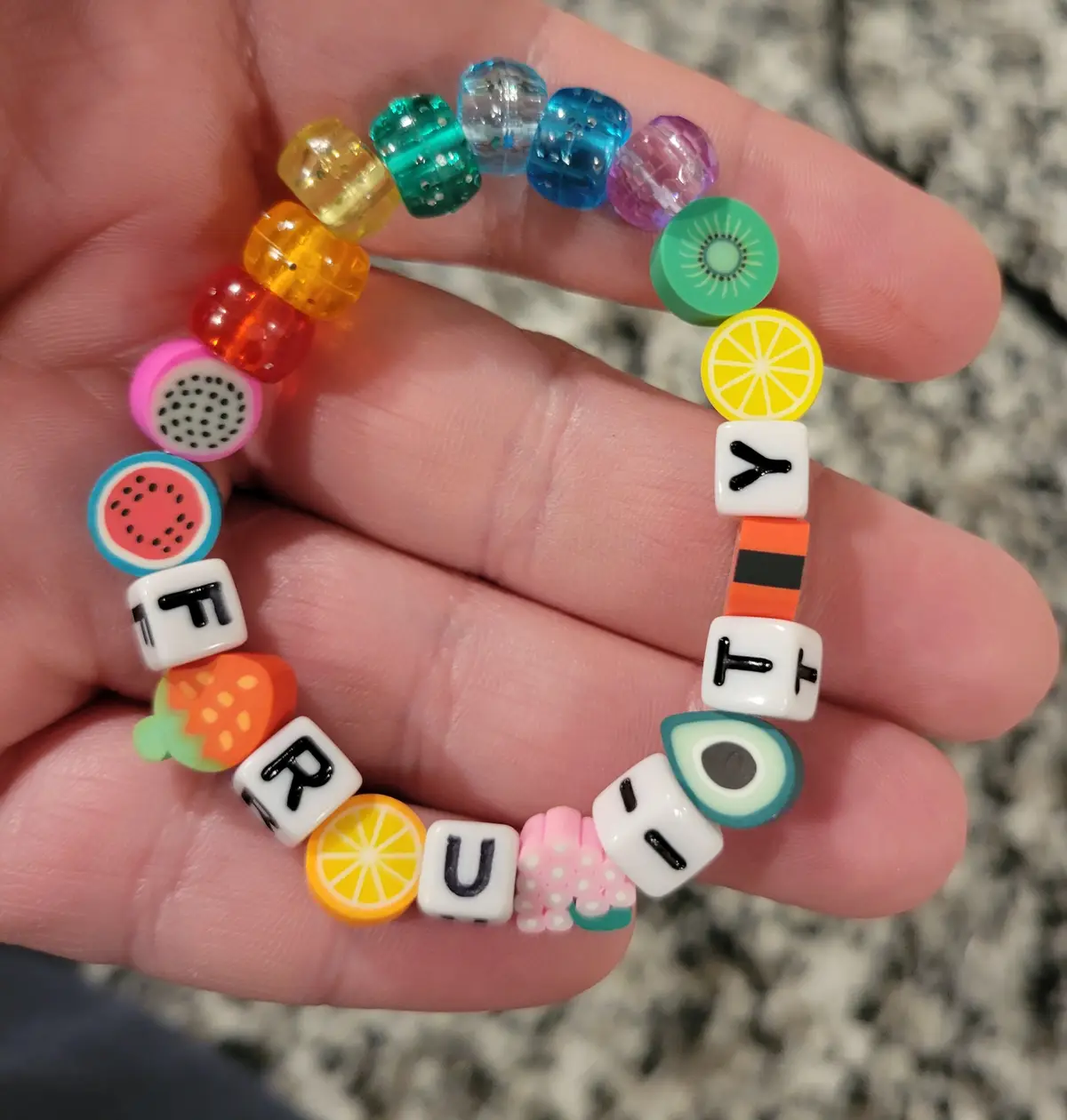 A hand holds a colorful beaded bracelet with various fruit-themed and letter beads spelling out 'fruit'. The beads include…