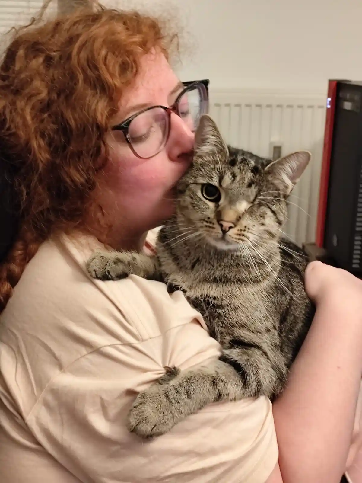 A person with curly red hair and glasses is kissing a gray tabby cat held in their arms. The cat looks directly at the…