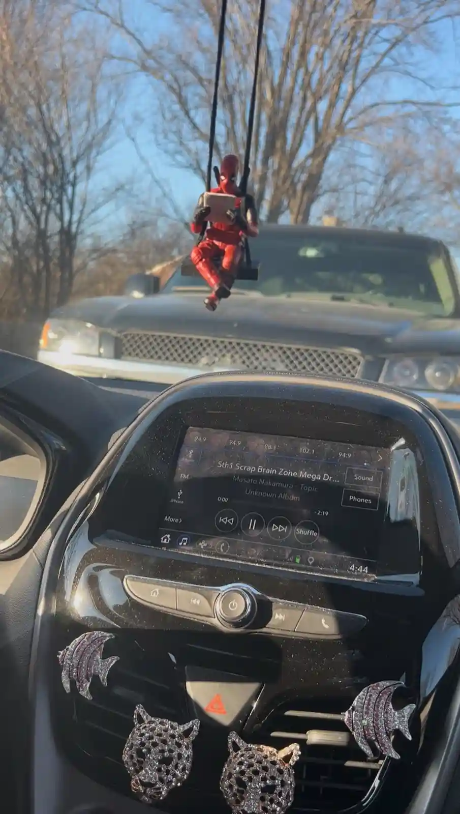A Deadpool action figure is suspended from a car's rearview mirror by a swing, holding a tablet as if working or browsing…
