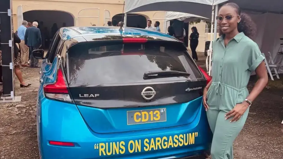A woman in a light green outfit stands smiling beside a bright blue Nissan Leaf electric car. The car's rear bumper has…
