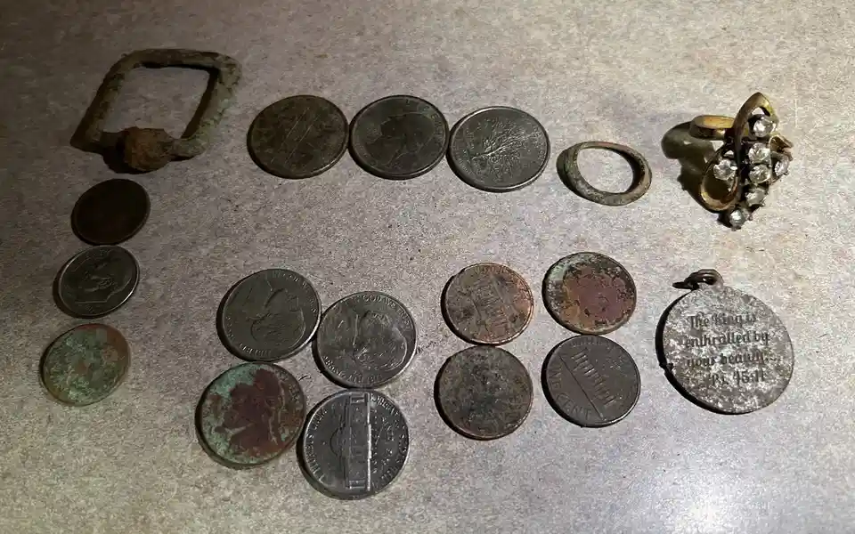 A collection of old coins, a rusty belt buckle, a simple ring, and a jeweled ring are laid out on a textured surface. Among…