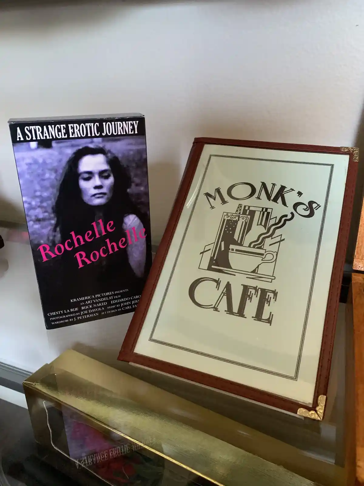 A VHS tape titled 'A Strange Erotic Journey' starring Rochelle Rochelle sits next to a framed 'Monk's Cafe' menu on a glass…